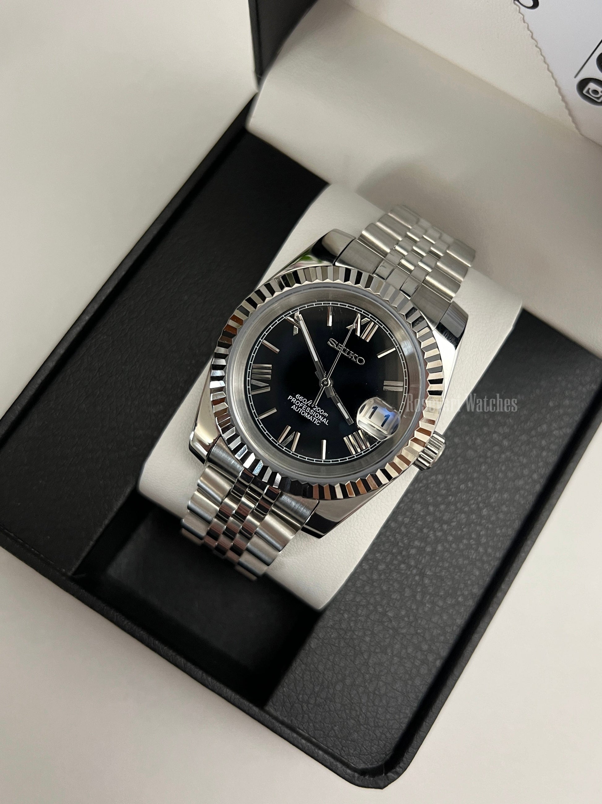 Datejust (Black Roman) Seiko NH35 automatic mod watch. A black dial watch with a silver fluted bezel and jubilee bracelet inside a box in a high angle shot.