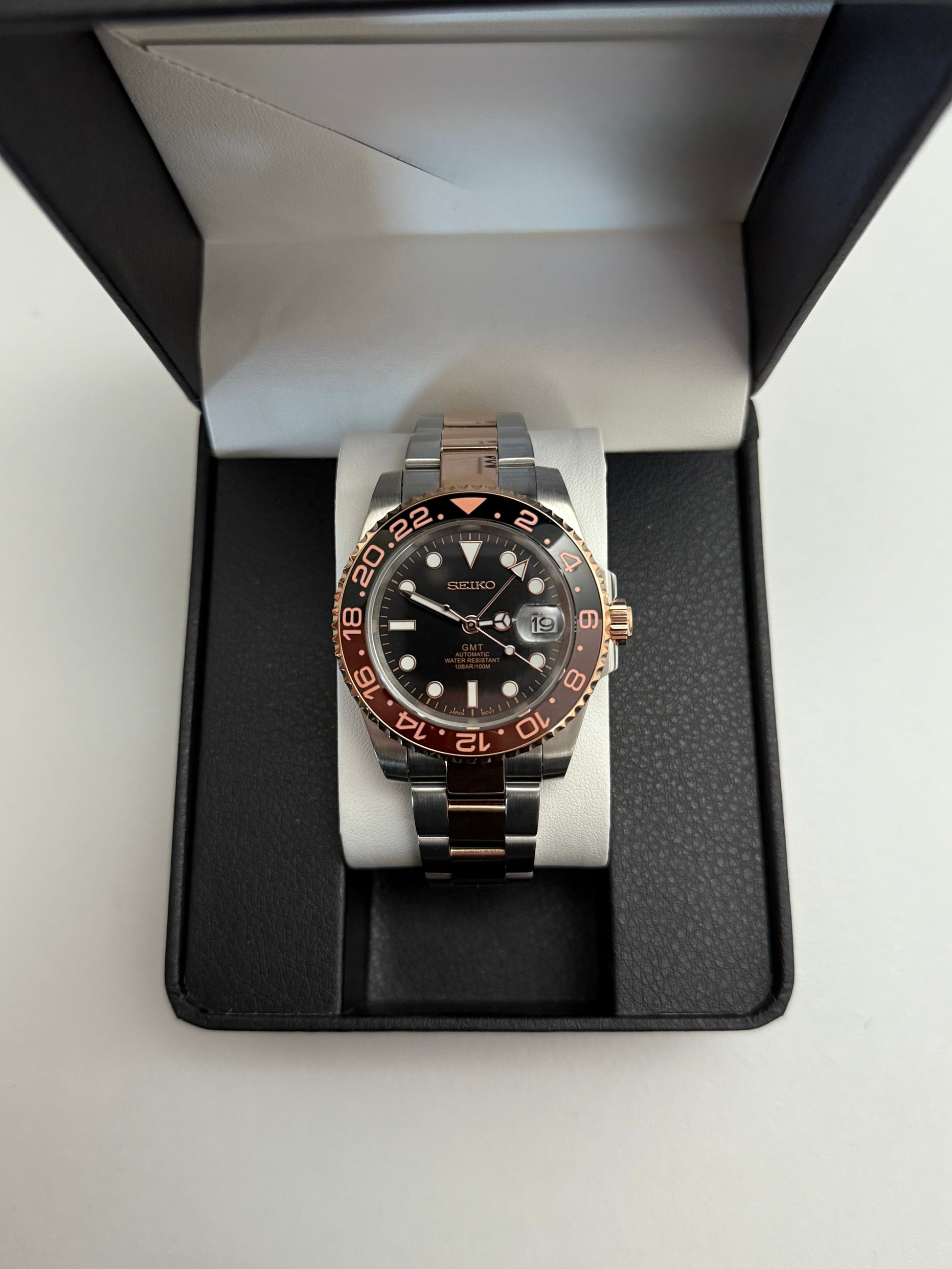 GMT (Rootbeer) Seiko NH34 GMT automatic mod watch. A full watch with a black dial and a black and rose gold bezel on a two tone oyster bracelet in a box.