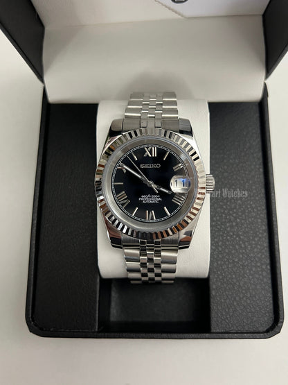 Datejust (Black Roman) Seiko NH35 automatic mod watch. A black dial watch with a silver fluted bezel and jubilee bracelet inside a box in a high angle shot.